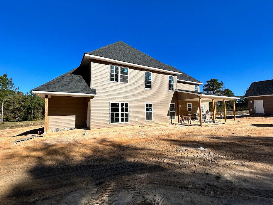 Partially built house with multi-pane windows, covered patio, exposed framing, surrounded by dirt and trees under a clear blue sky