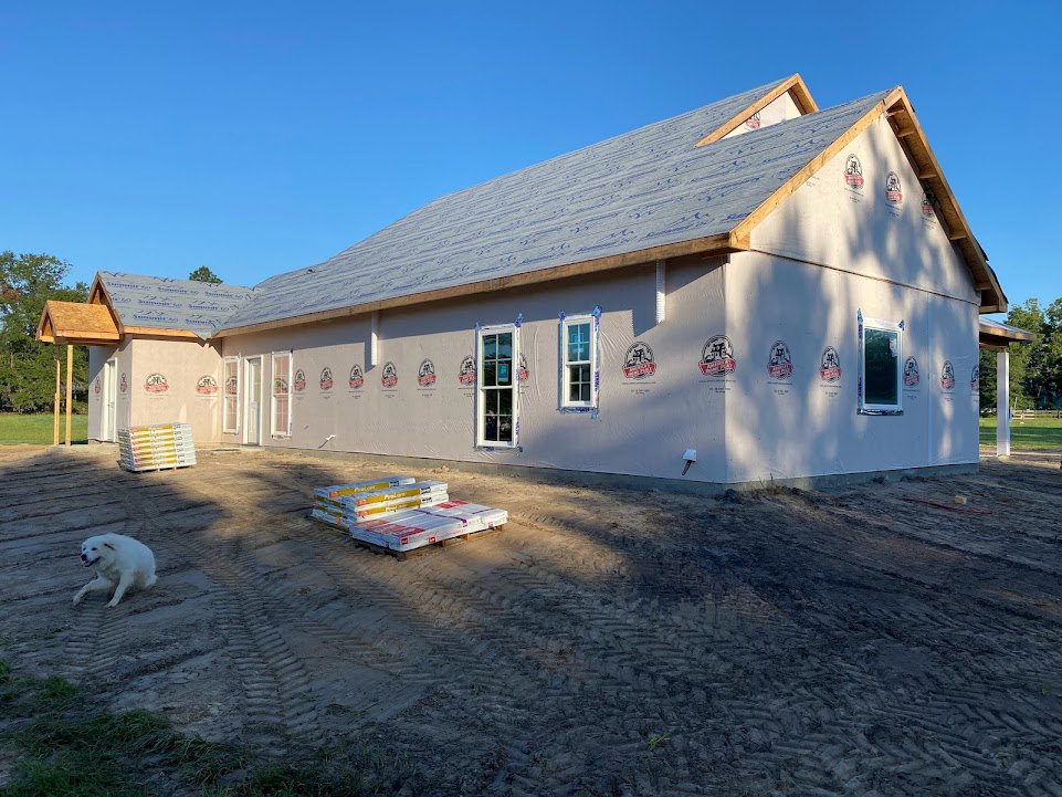 Partially built house with exposed framing, stacks of white and yellow bricks, plastic containers, and wooden pallets on dirt ground; white dog running along dirt road under clear
