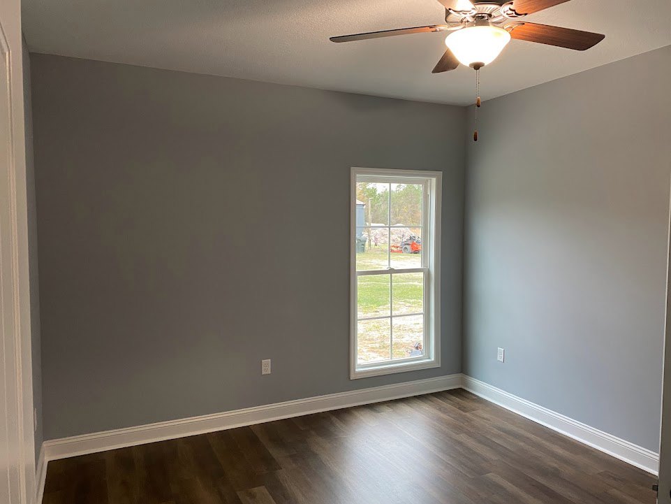 Wood floor room with white plaster walls, ceiling fan with light fixture, large window overlooking field, trees, and distant tractor.