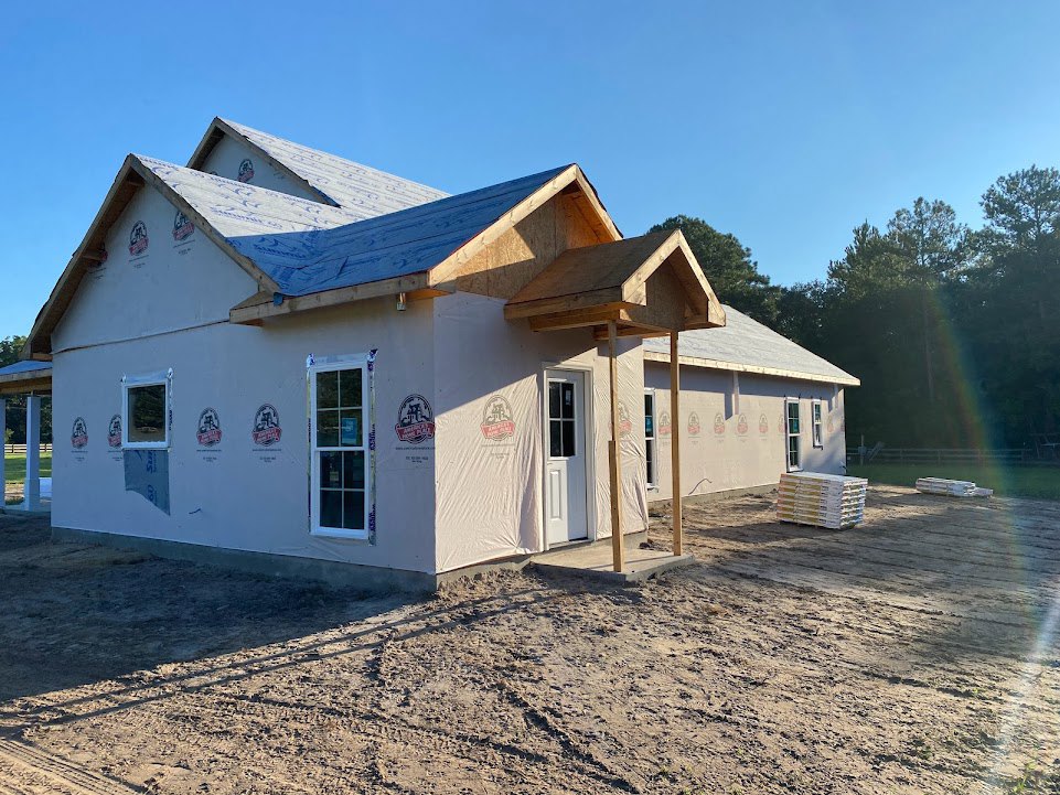 Two-story house under construction with plastic sheeting, glass door displaying a sign, white door with window, dirt yard, and stacked pallets of paper in front