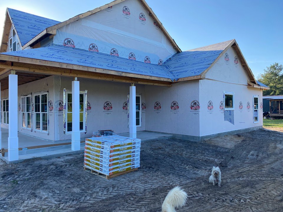 Partially built house with exposed framing, stack of wooden pallets on dirt ground, white long-haired dog with fluffy tail near construction materials, windows and roof visible