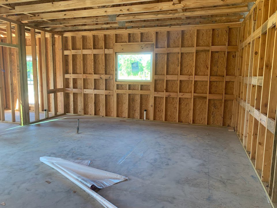 Unfinished room with exposed wood ceiling beams, concrete floor, roll of plastic sheeting on the ground, window with a paper sign attached