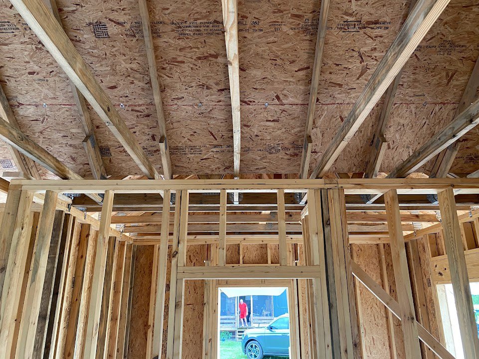Exposed wood framing and ceiling with black text, unfinished interior walls, a man in red shorts walking through a glass door, another man in a red shirt standing on the porch