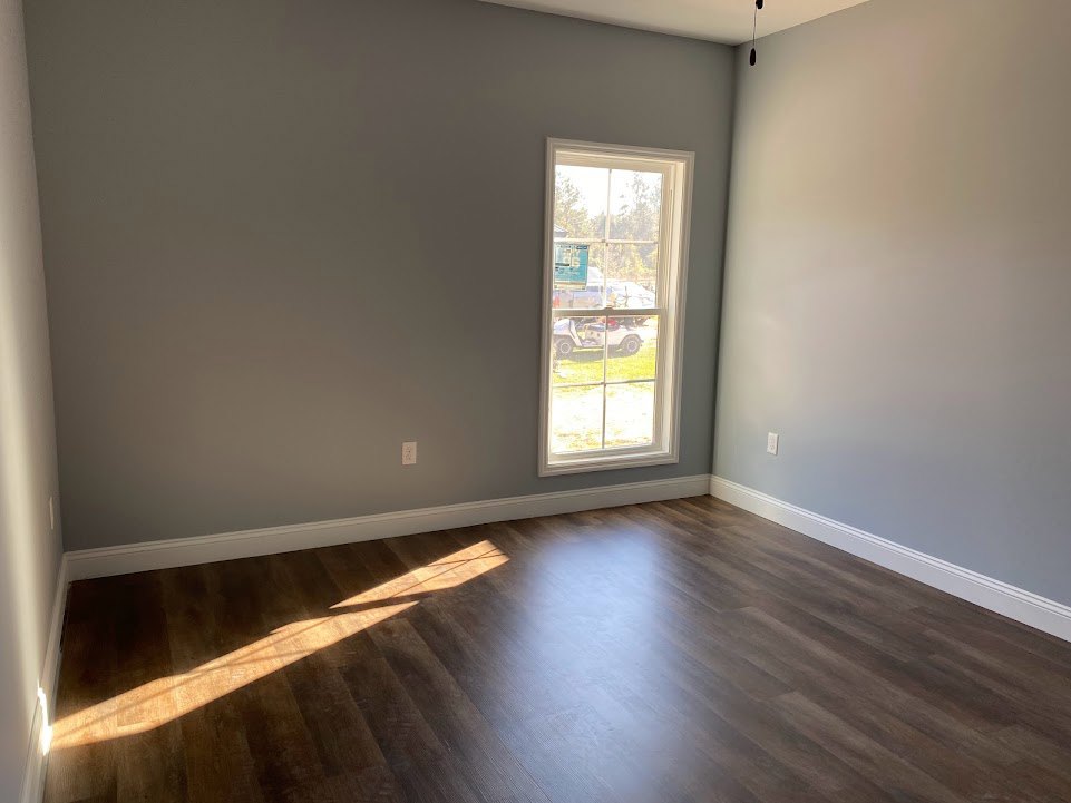 Sunlit hardwood floor in a room with white walls and a large window overlooking a parked white car.