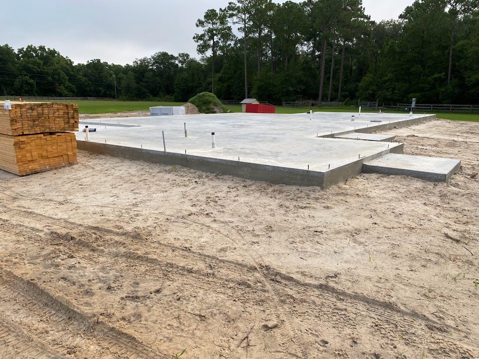 Concrete foundation surrounded by sand, stacks of wood planks, red utility box, and dense trees under a blue sky with clouds