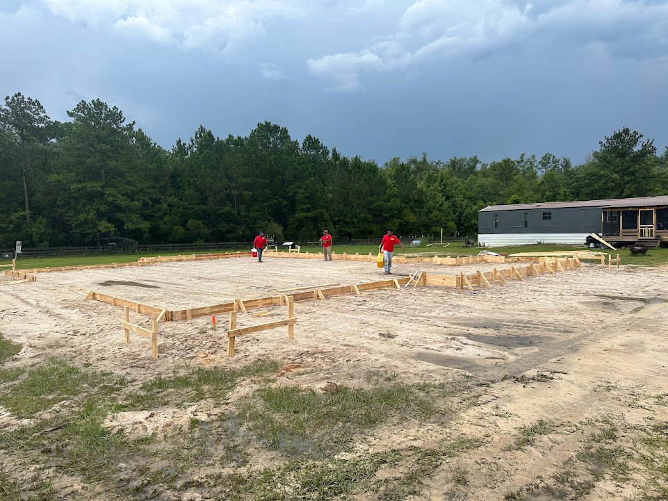 Construction crew in red shirts standing on dirt lot near parked trailer, unfinished building, and surrounding trees under cloudy sky