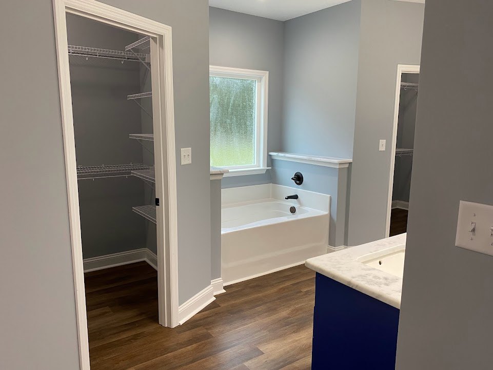 White freestanding bathtub on wood plank flooring, frosted glass window, light switch on wall, open closet with built-in shelves