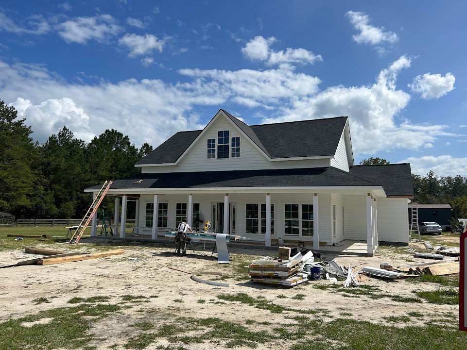 Partially built house with exposed framing, roof shingles, and windows; two ladders leaning against the exterior; man standing near porch; stack of pallets and box in foreground