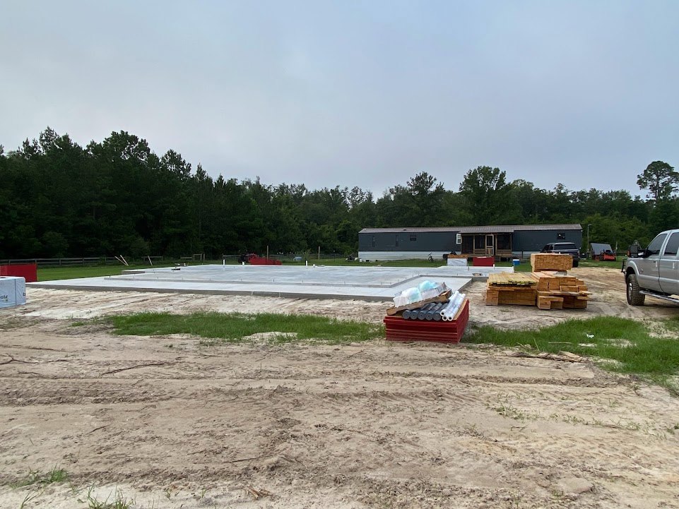 Concrete foundation slab surrounded by dirt, metal pipes, and construction materials; rolls of plastic stacked on a red box; parked truck and van near site; partially built house