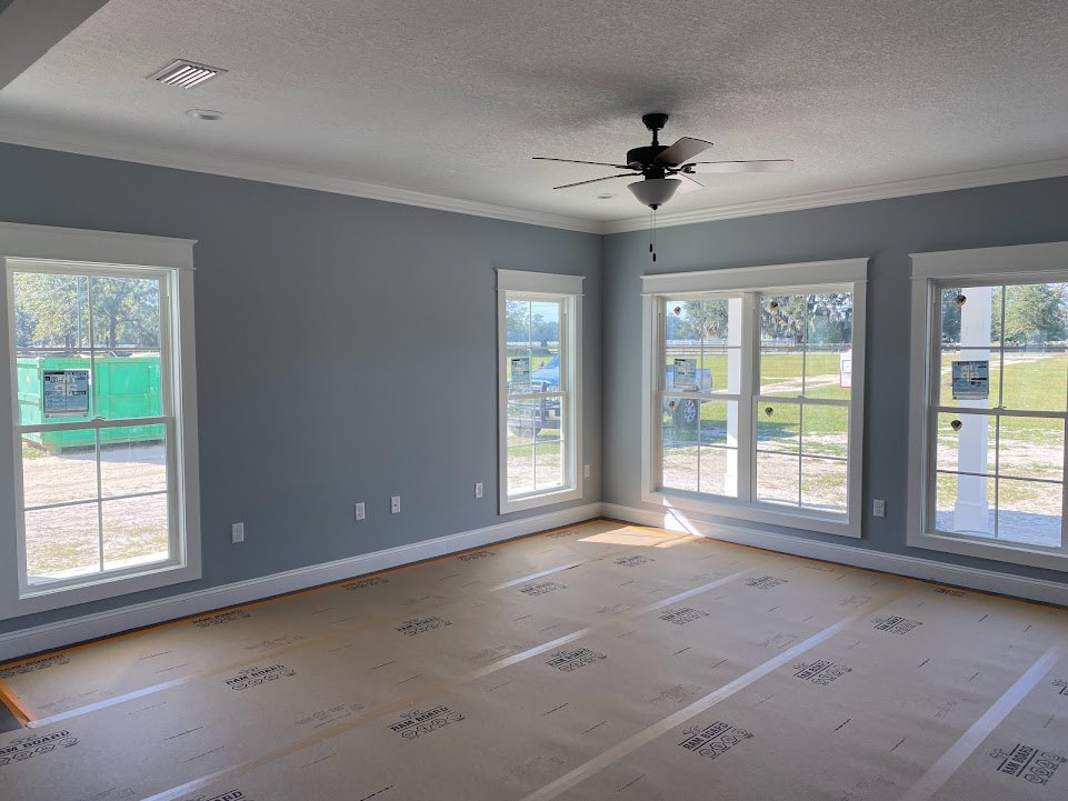 Bright room with multiple windows, ceiling fan with light fixture, cardboard covering part of the floor, white door, view of grass field and green container outside