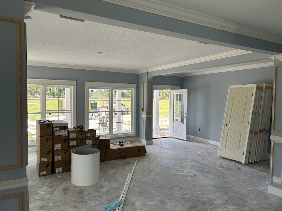 Unpacked cardboard boxes stacked near a white door with glass window, white framed window, light-colored plaster walls, and neutral flooring in a residential room.