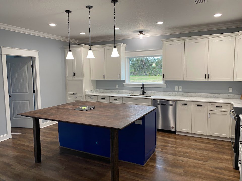 Spacious kitchen featuring a large white island with black handles, white cabinetry, a person loading a dishwasher, a window with a green lamp and view of trees, and a table with a