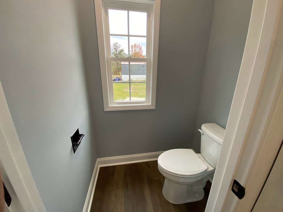 White toilet beneath a window overlooking grass and trees, light-colored tile floor, neutral walls, minimal bathroom accessories