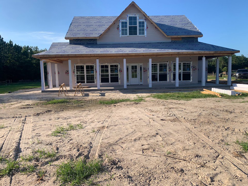 Partially built house with shingled roof, multi-pane windows, dirt yard marked by tire tracks, scattered grass patches, and wooden porch supports.