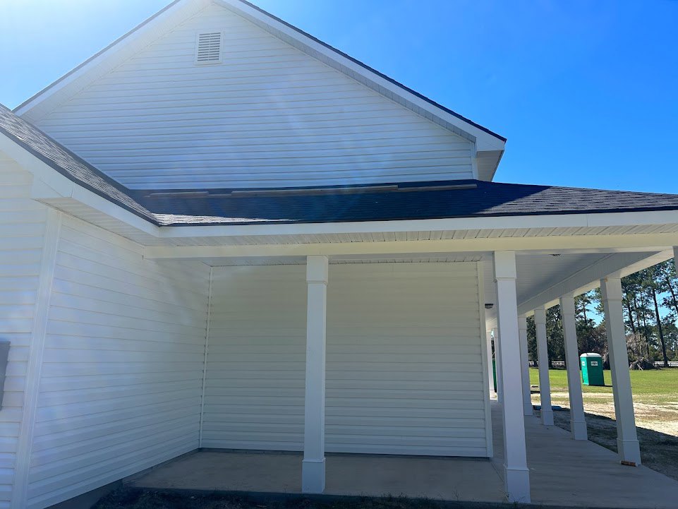 White house exterior with white pillars on a covered porch, gray shingle roof, white siding, windows, green lawn, and a person walking on the sidewalk