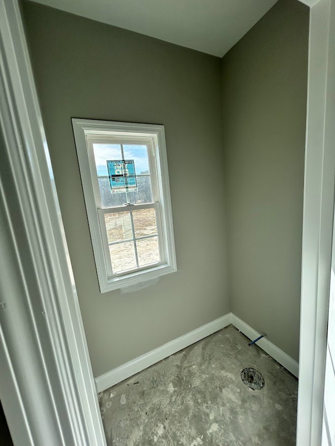 Room with large window, unfinished concrete floor, white plaster walls, and ceiling; sunlight streaming in, construction sign visible through window.