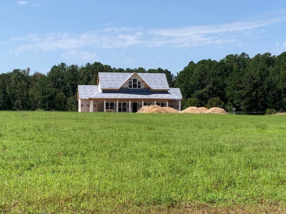Single-story house with triangular roof, piles of sand in front, large grassy field, scattered trees, and blue sky with clouds