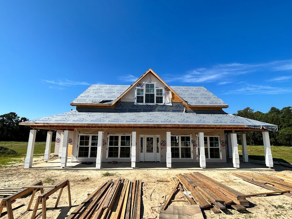 Wood-framed house under construction with exposed beams, stacked lumber on sandy ground, glass-paneled white door, multiple windows, and blue sky overhead