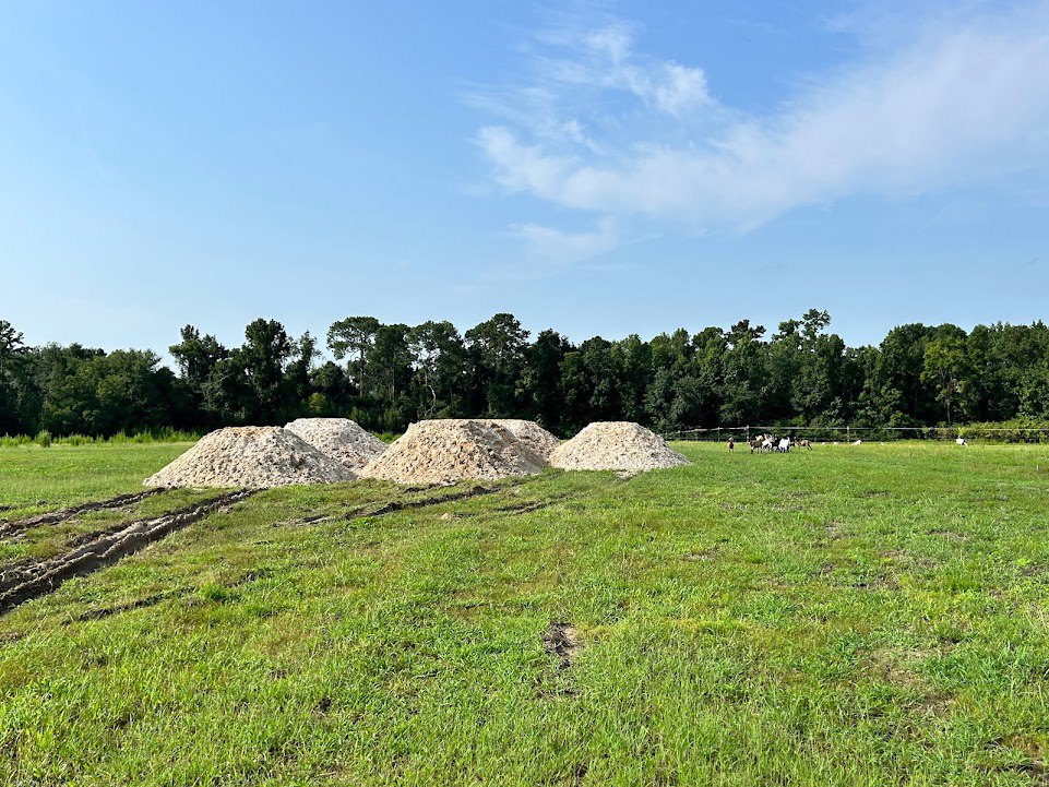 Several piles of gravel and dirt scattered across an open grassy field, with a backdrop of trees under a partly cloudy blue sky.