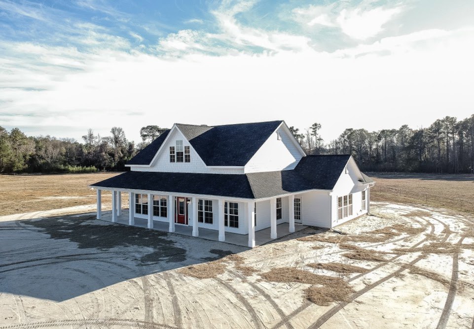 White two-story house with wide covered porch, red front door, multiple windows, concrete driveway, and blue sky with scattered clouds