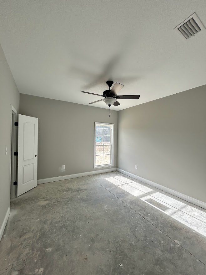Neutral-toned room with concrete floor, white walls, ceiling fan with light fixture, window displaying a sign, ceiling vent, and white door with black handle