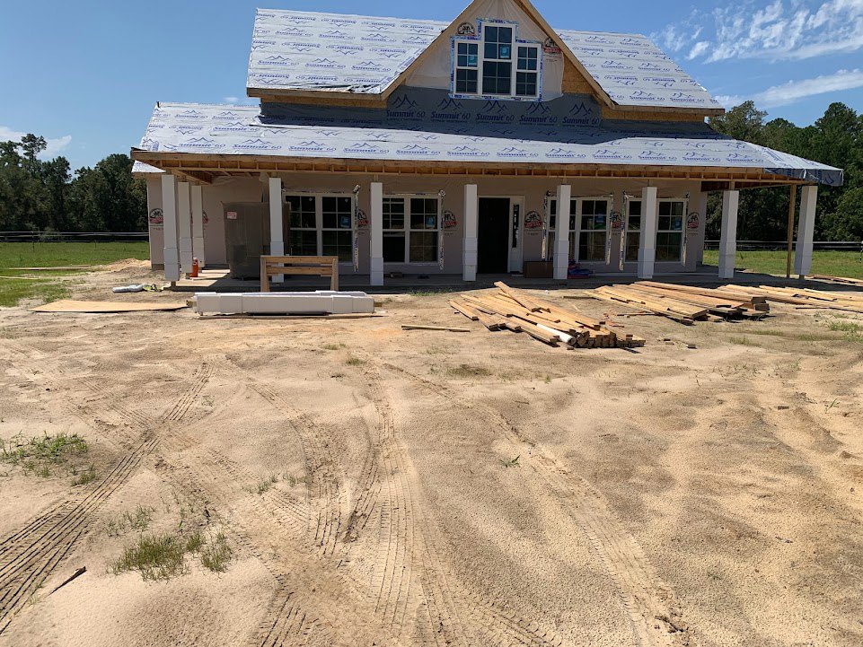 Framed house under construction with exposed wooden beams, unfinished roof, piles of lumber on ground, and window with temporary signage