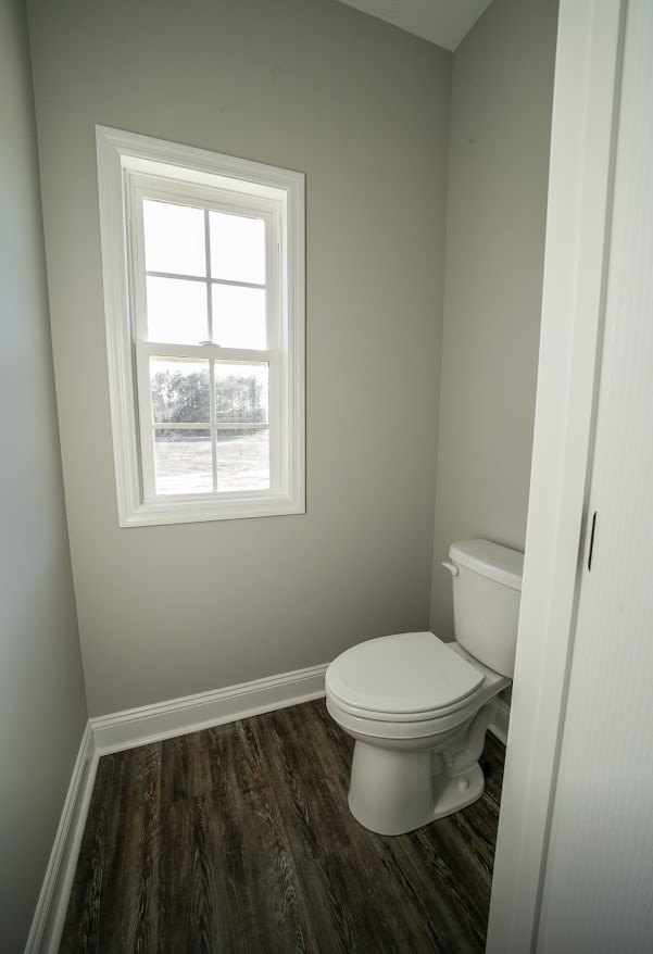 White toilet with closed lid on wood floor, small bathroom with white window and door, light-colored walls, minimal decor.