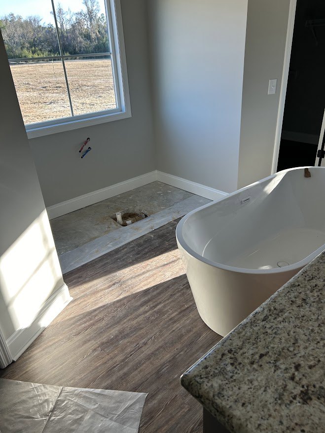 White freestanding bathtub beside a wide window overlooking grassy field, light wood vanity with undermount sink, neutral tile floor, chrome fixtures, plastic bag on countertop