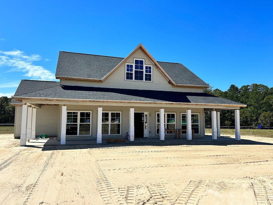 Modern house with multiple large windows, black front door with white trim, tire tracks on sandy dirt road leading to entry, blue sky overhead, porch area visible, surrounded by