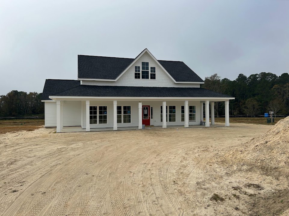 White siding house with red front door, multiple windows, columned entry, dirt driveway with tire tracks, blue utility box, surrounded by trees.