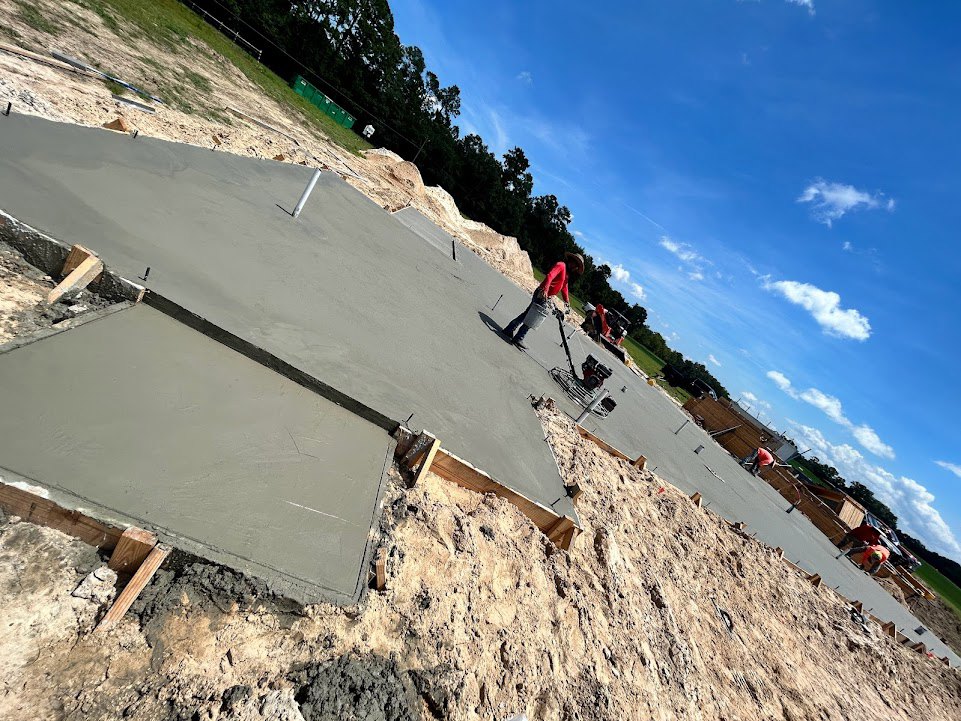 Worker in red shirt carrying a bucket across unfinished concrete slab on residential construction site, dirt area and cloudy sky in background