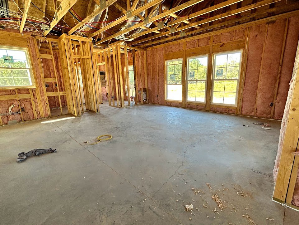 Living room with exposed wood ceiling beams, large windows overlooking trees, hardwood floors, and a yellow hose coiled near the wall.