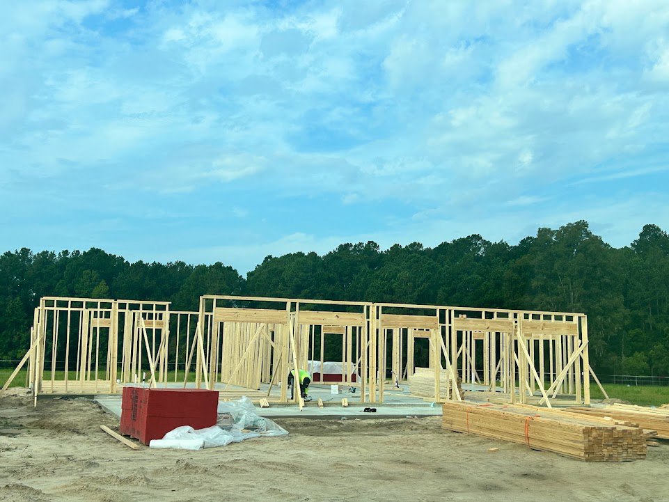 Wood-framed house under construction with exposed beams, stacks of lumber, worker on site, and trees in the background under a partly cloudy blue sky