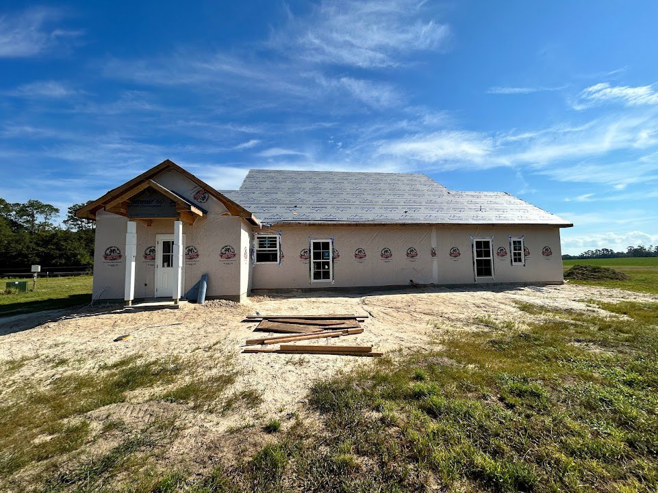 Framed house under construction with exposed wooden roof trusses, pile of lumber on grassy lot, clear blue sky overhead