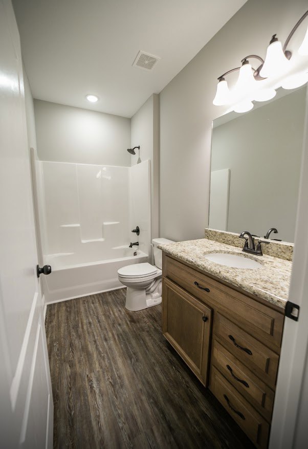 Bathroom with marble countertop sink, wood cabinetry, wall-mounted mirror, toilet, and wood-grain tile flooring