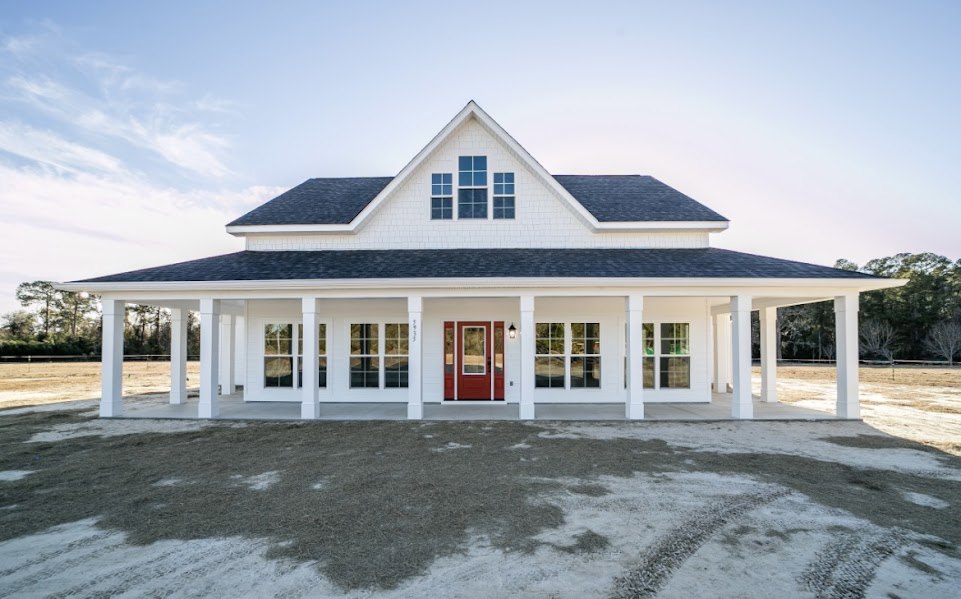 White siding house with prominent red front door, white trim, covered porch, triangular window in gabled roof, green lawn, and trees under partly cloudy sky