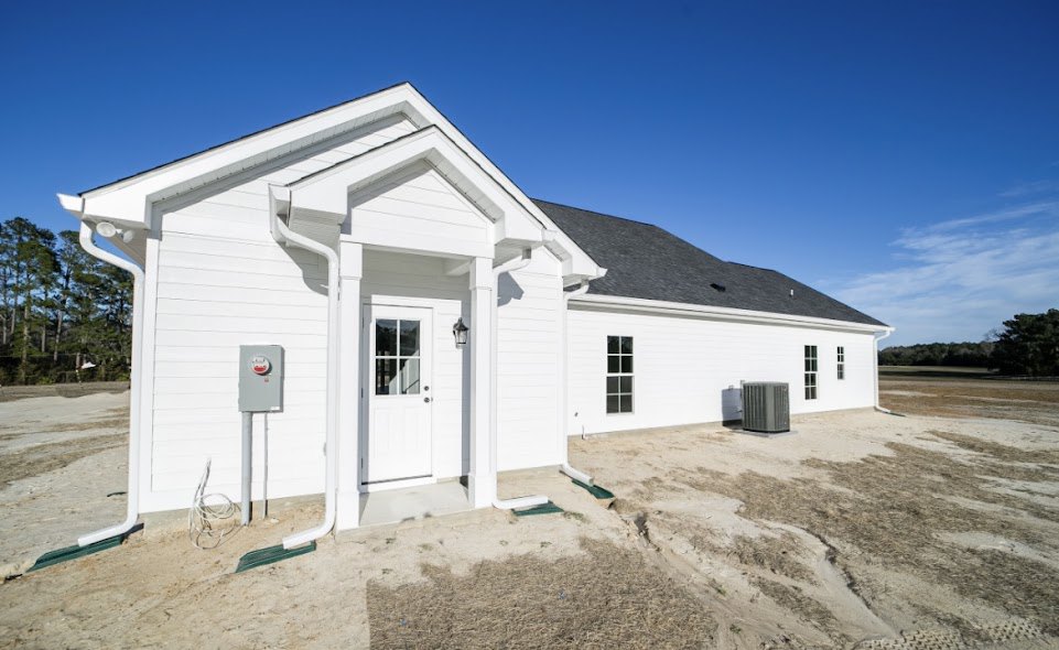 White two-story house with horizontal siding, dark roof, front door, and multiple windows, set against a clear blue sky with scattered clouds and surrounded by green lawn and