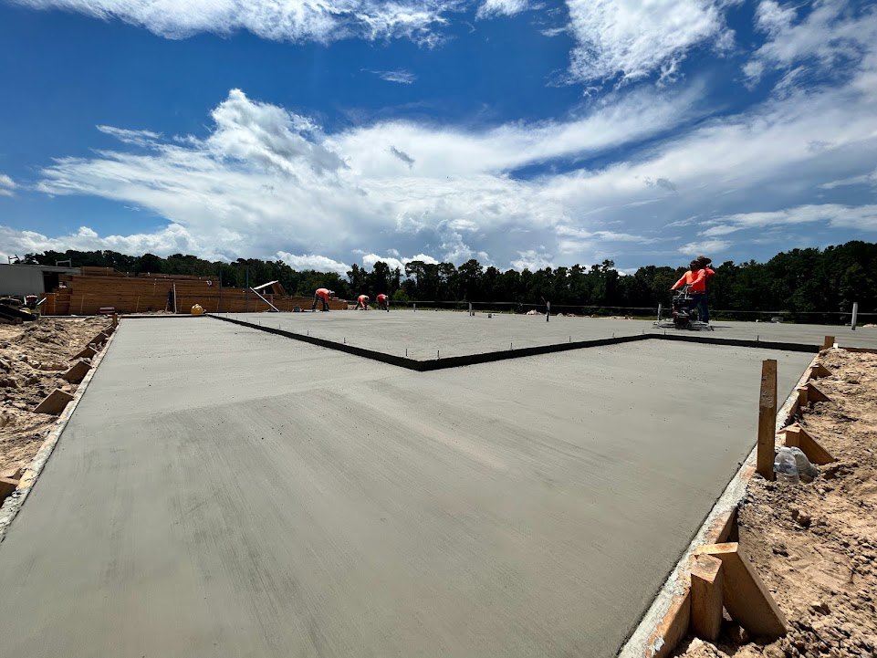 Freshly poured concrete slab surrounded by sand and dirt, group of men working in the background under a blue sky with scattered white clouds, trees lining the horizon.