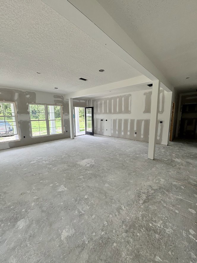 Open white door leading to a bright room with large windows, white plaster walls, concrete flooring, and a car visible outside through the window.