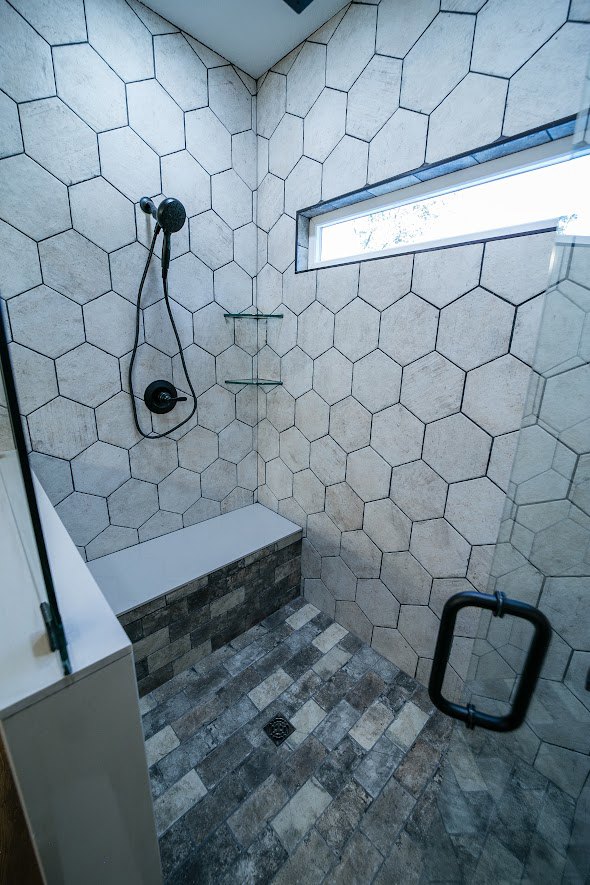 Walk-in shower with chrome shower head, white tile walls, window, black wire on tile surface, white cabinet with black handle, rectangular wall light fixture, white tile floor with