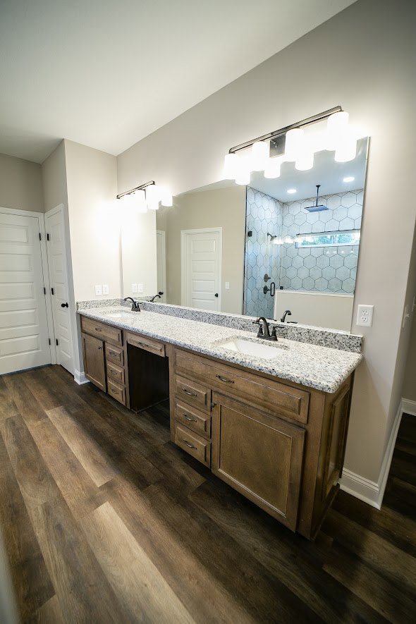 Bathroom featuring a wide framed mirror above a double sink vanity with white cabinetry, black hardware, and light tile flooring; white door with black handle visible on the side.