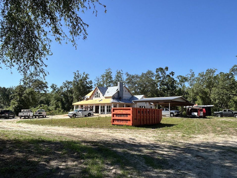 Modern house under construction with white siding, large red-orange shipping container in front, white truck parked on dirt driveway, grassy lot bordered by trees under clear blue