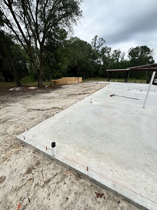 Concrete slab with metal poles, picnic table under metal structure, trees and bench in background, cloudy sky overhead