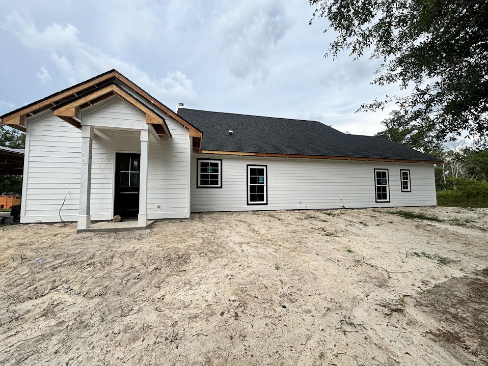 White siding house with black shingle roof, black front door featuring a white square window, dirt yard, large window with a sign, cloudy sky above.