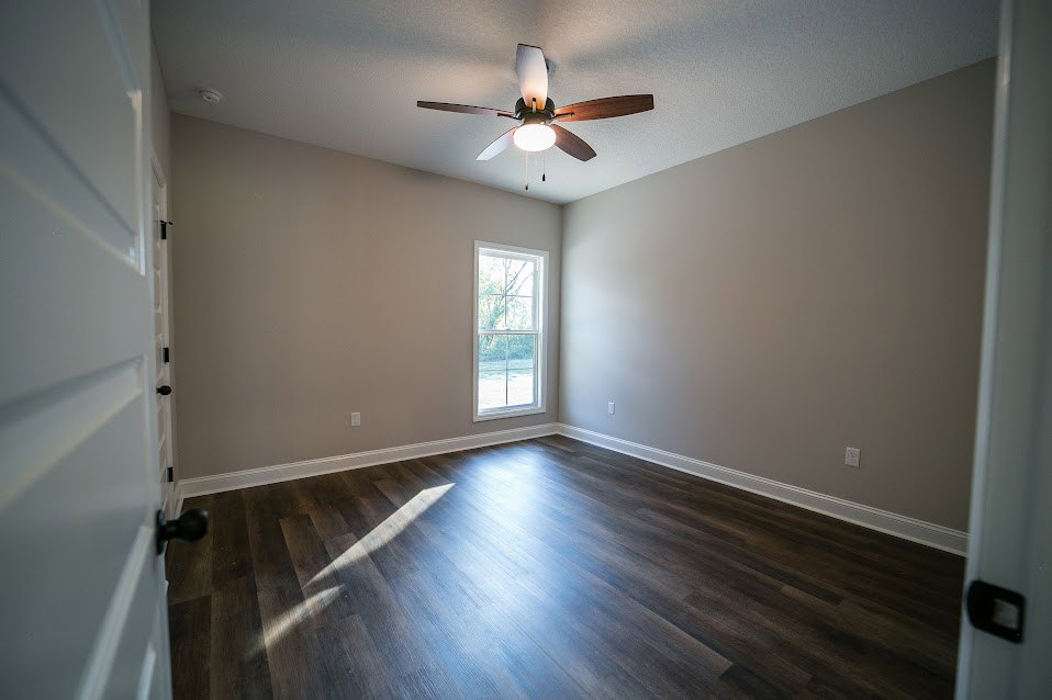 Dark wood flooring, white walls, ceiling fan with light fixture, white-framed window, white door with black hardware