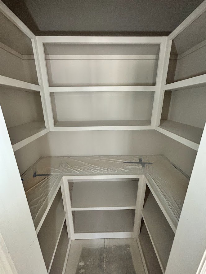 White built-in shelves line one wall of a closet-style room with a matte black ceiling and plaster walls; some shelves are covered with plastic sheeting and one features a black