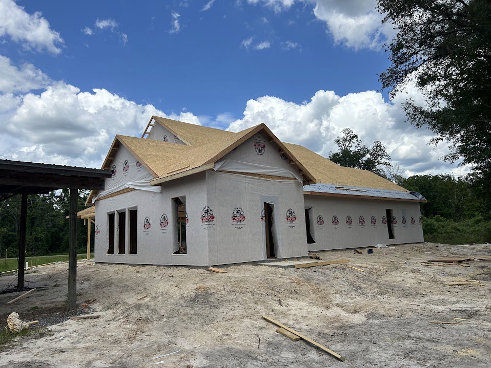 Wood-framed house under construction with exposed beams, unfinished roof, scattered wood planks and rocks on dirt lot, surrounded by trees beneath a clear blue sky
