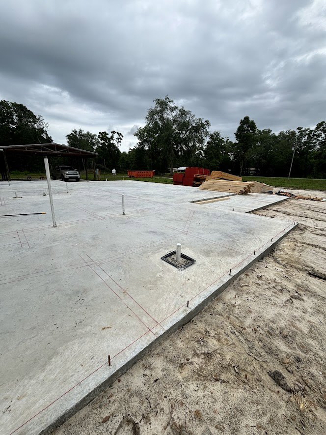 Concrete slab foundation with stacked lumber and a parked car, surrounded by trees under a cloudy sky