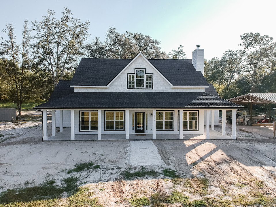 White two-story house with black roof and door, expansive concrete driveway, multiple large windows, small porch, and mature trees in the yard.