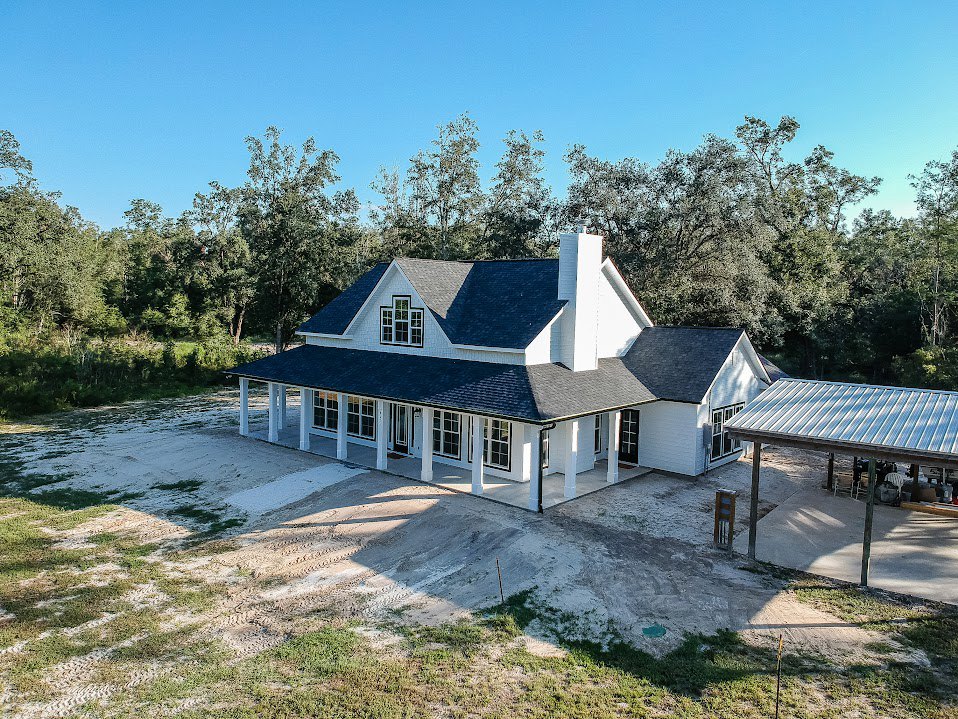 White house with black roof, covered patio with shade, dirt path leading to entrance, surrounded by trees under blue sky